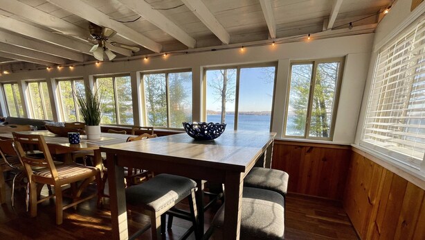 Un comedor con mesa y sillas de madera, ventanas amplias con vistas al exterior y un ventilador de techo.