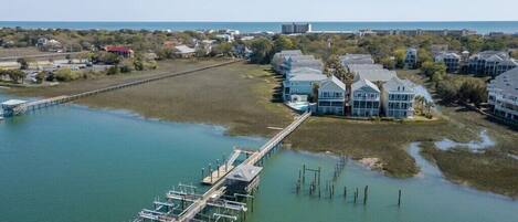 An aerial view of a coastal property with a dock extending into a calm body of water, surrounded by residential buildings and a clear view of the ocean.