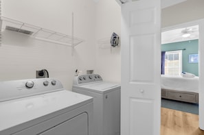 A laundry room with two front-loading washers and dryers, a drying rack, and a door leading to a bedroom with a bed and a window. - Sea Glass at Waters Edge | Riverfront 3BR/3BA Townhome, Pool, Pier & Walk to Folly Beach (Folly Beach)