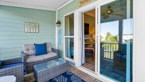 Un porche cubierto con muebles de mimbre, una puerta de cristal que da a una terraza, y un cartel que dice 'BEACH'.