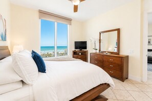 A hotel room with a bed, a TV, a dresser, a mirror, and a view of the ocean.