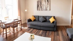 Una sala de estar con un sofá gris, sillas de madera, una mesa de centro blanca con flores y un cuadro en la pared.