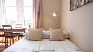 Una cama bien hecha con dos almohadas y dos toallas dobladas, un escritorio con sillas y una lámpara en la pared.