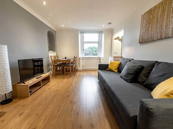 A modern living room with a grey sofa, wooden dining table, and a TV on a stand. - ☆Bright Apartment, Near University + City Centre☆ (Dundee)