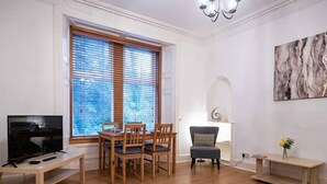 Un salon comprenant une table à manger en bois, des chaises, un téléviseur et une grande fenêtre avec des stores.