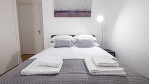 Un lit bien fait, avec une couverture grise et des oreillers blancs, une lampe de chevet et un tableau accroché au mur.