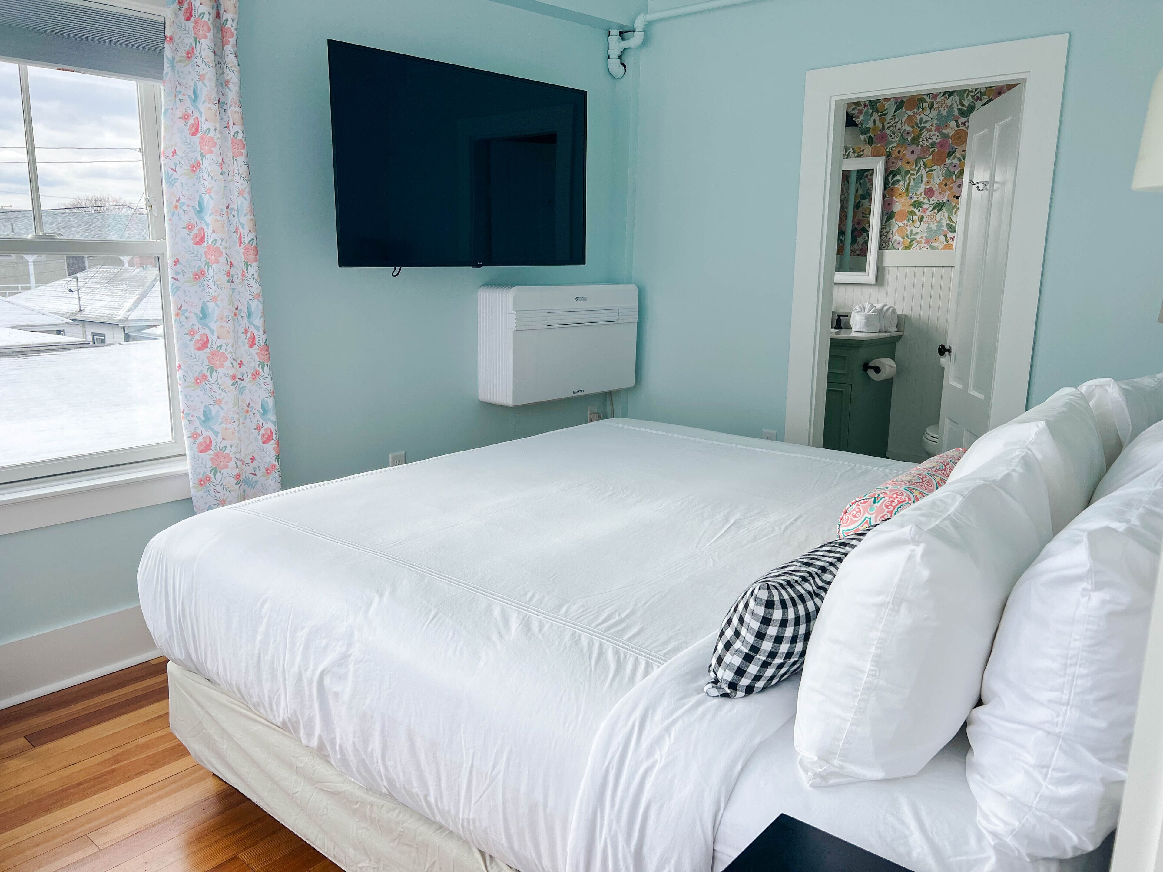 A bedroom with a large bed, a mounted TV, an air conditioning unit, and a window with floral curtains.