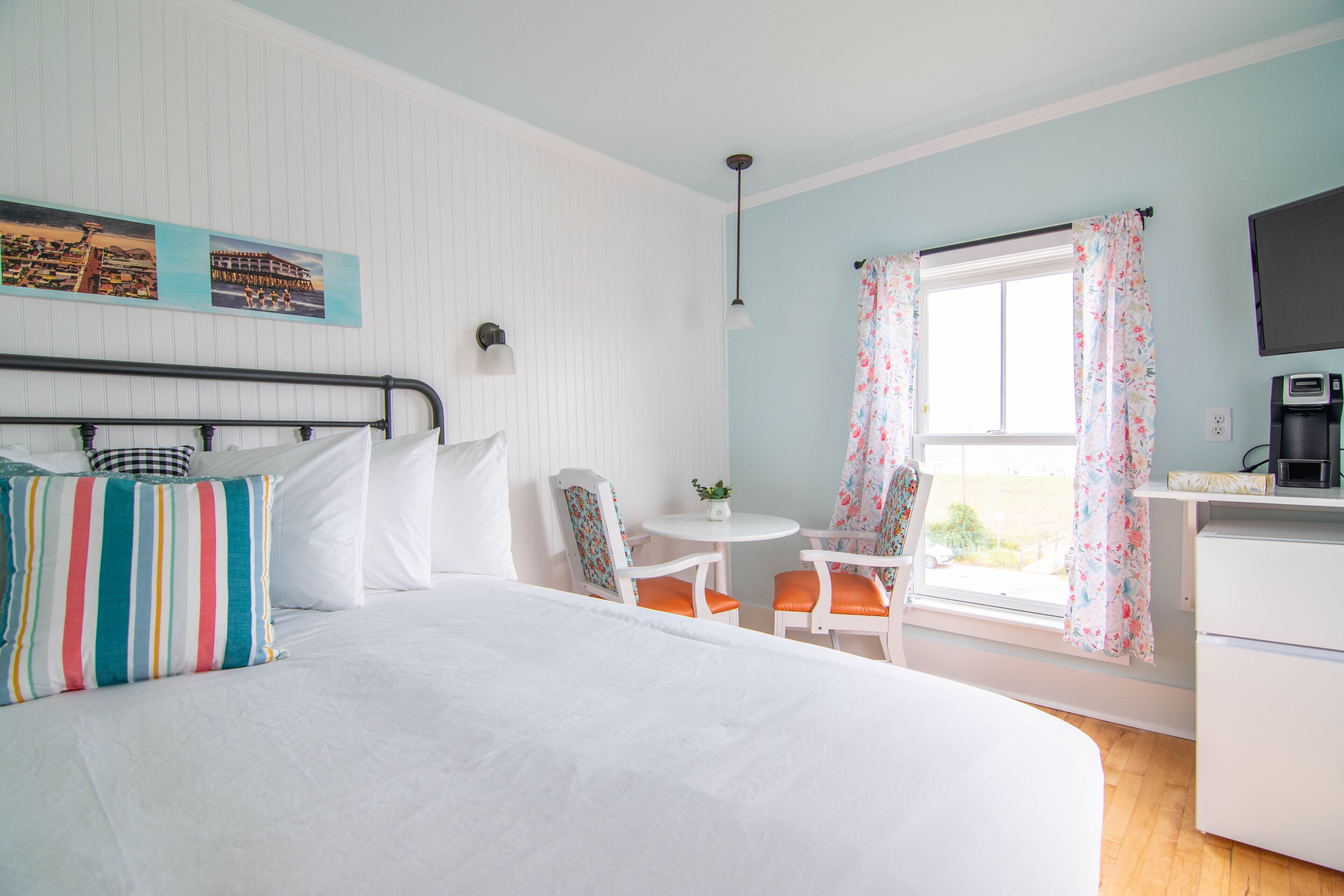 Une chambre à coucher comprenant un lit, un bureau, une chaise, une petite table, un téléviseur et une fenêtre avec des rideaux.