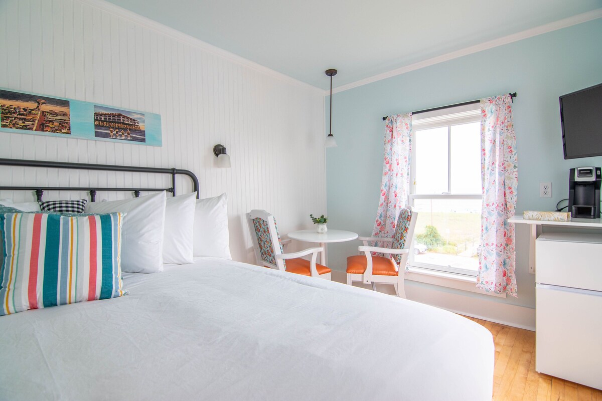Ein Schlafzimmer mit Bett, Schreibtisch, Stuhl, kleinem Tisch, Fernseher und einem Fenster mit Vorhängen.