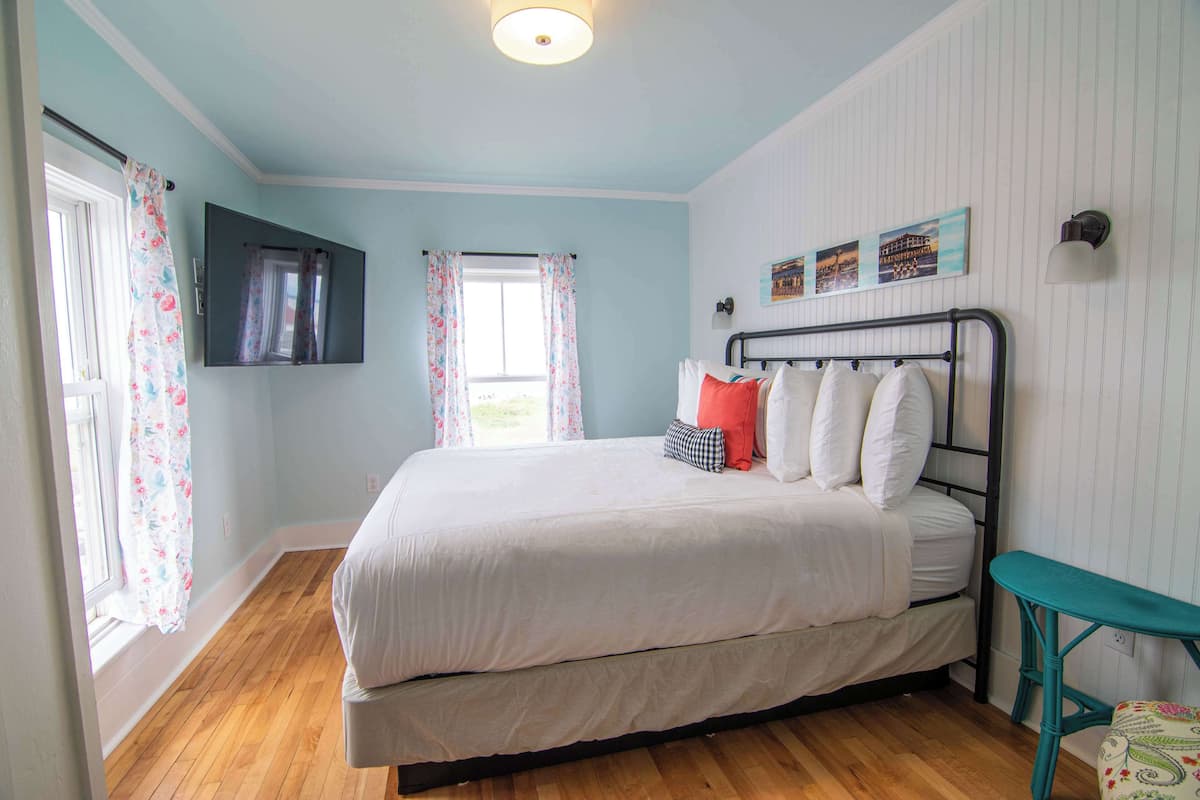 A bedroom with a bed, a window with curtains, a wall-mounted TV, and a small table.