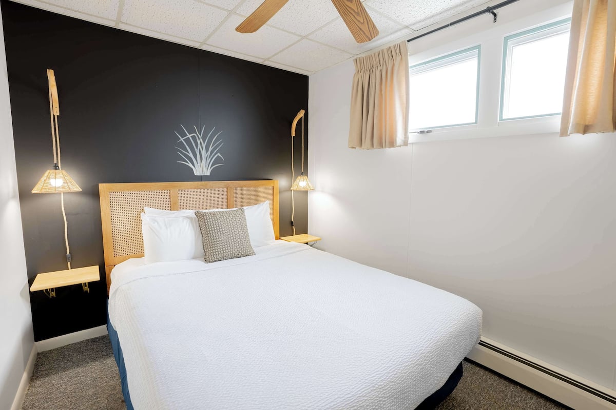 A neatly made bed with a white comforter and a wooden headboard. There are two bedside tables with lamps, and a wall-mounted shelf above the bed.