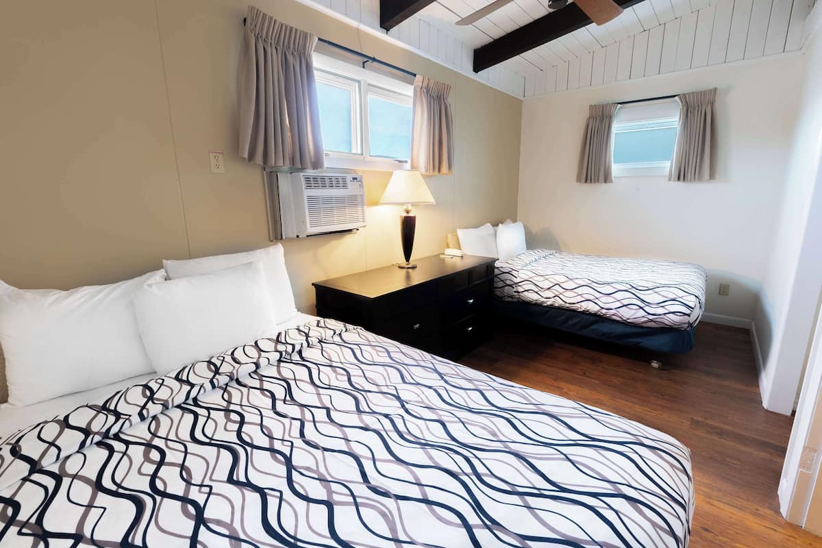 A bedroom with two beds, a bedside table with a lamp, an air conditioning unit, and a window with curtains.