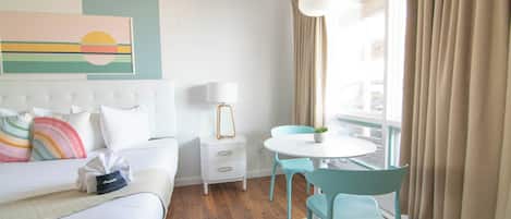 Habitación de hotel con cama, mesita de noche con lámpara, pequeña mesa con sillas y ventana con cortinas.