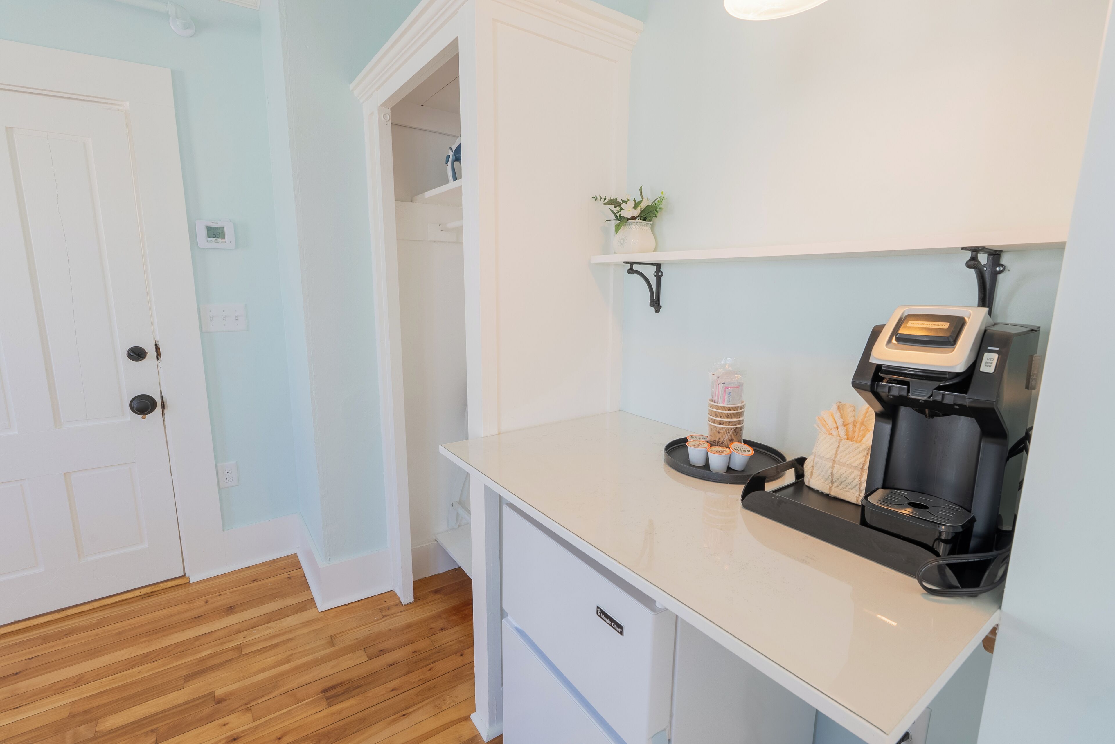 A kitchen with a coffee machine, a shelf with a plant, and a door leading to another room.