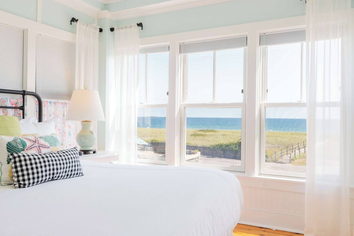 A bedroom with a large bed, a view of the ocean through multiple windows, and a lamp on the nightstand.
