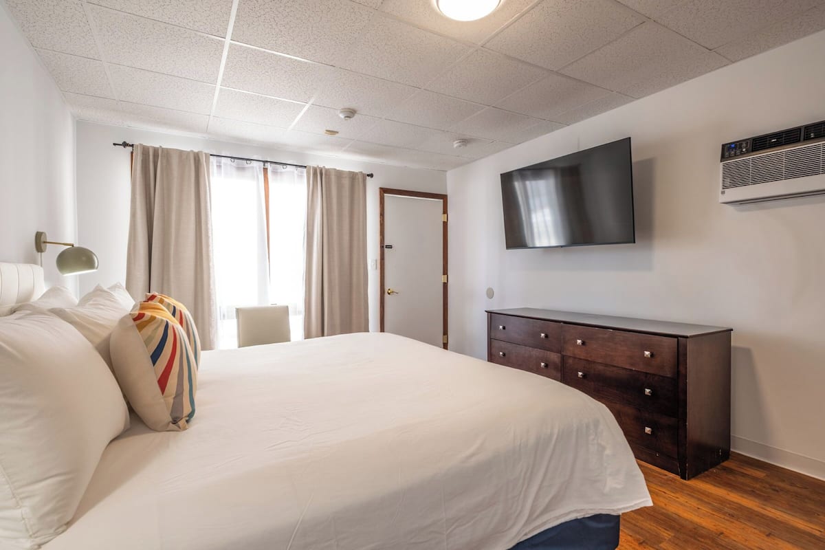 A hotel room with a large bed, a TV mounted on the wall, a dresser, and a window with curtains.