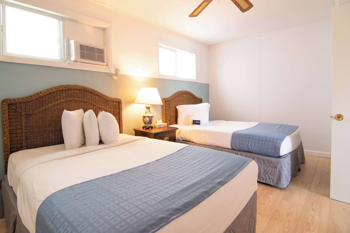 A hotel room with two beds, a wicker headboard, a bedside lamp, and a window with an air conditioning unit.