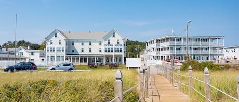 A coastal hotel with a wooden boardwalk leading to the entrance, featuring multiple stories and a balcony on the upper level.