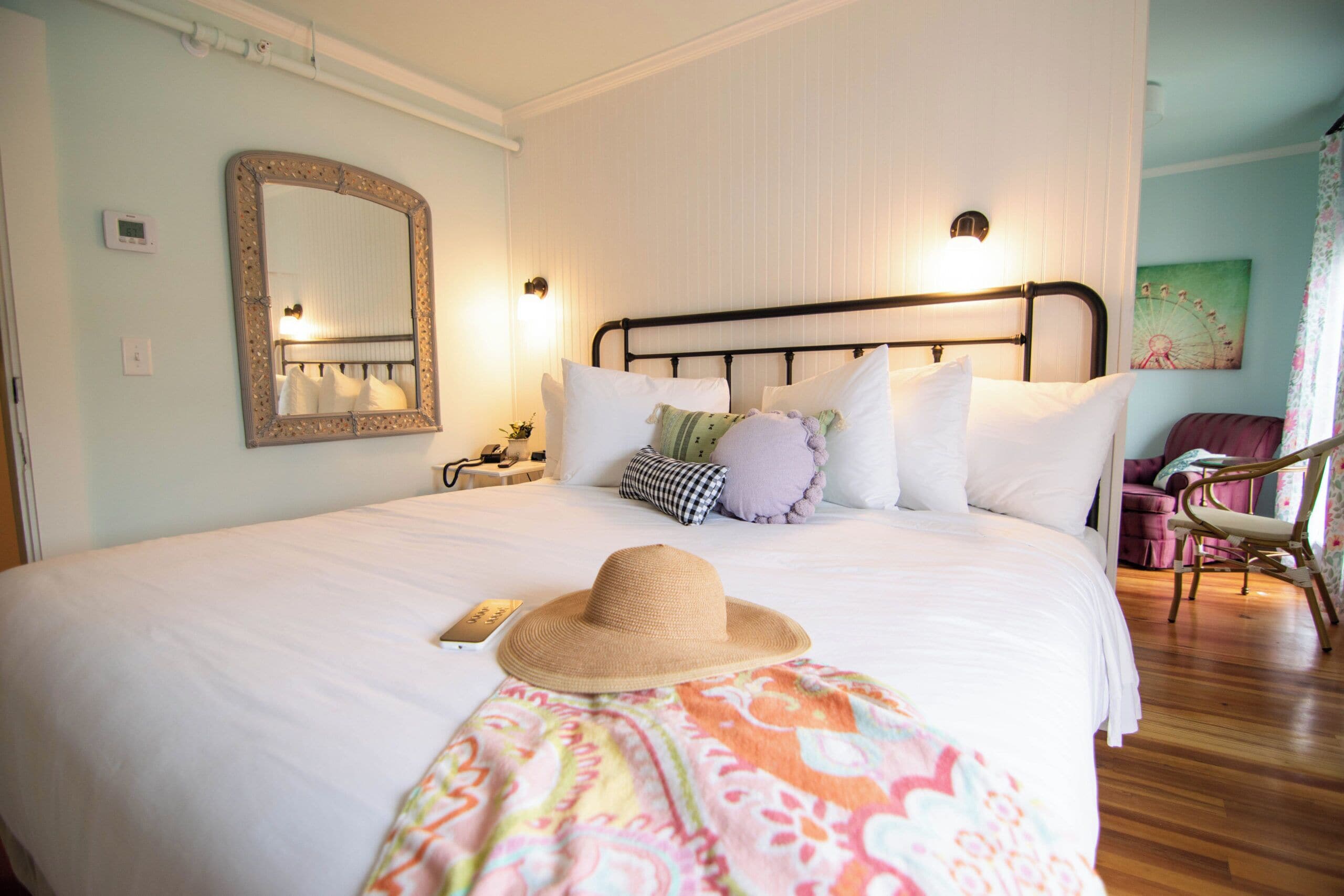 A neatly made bed with a hat and a colorful blanket, a bedside table with a lamp, and a chair in a room with light-colored walls and a large mirror.