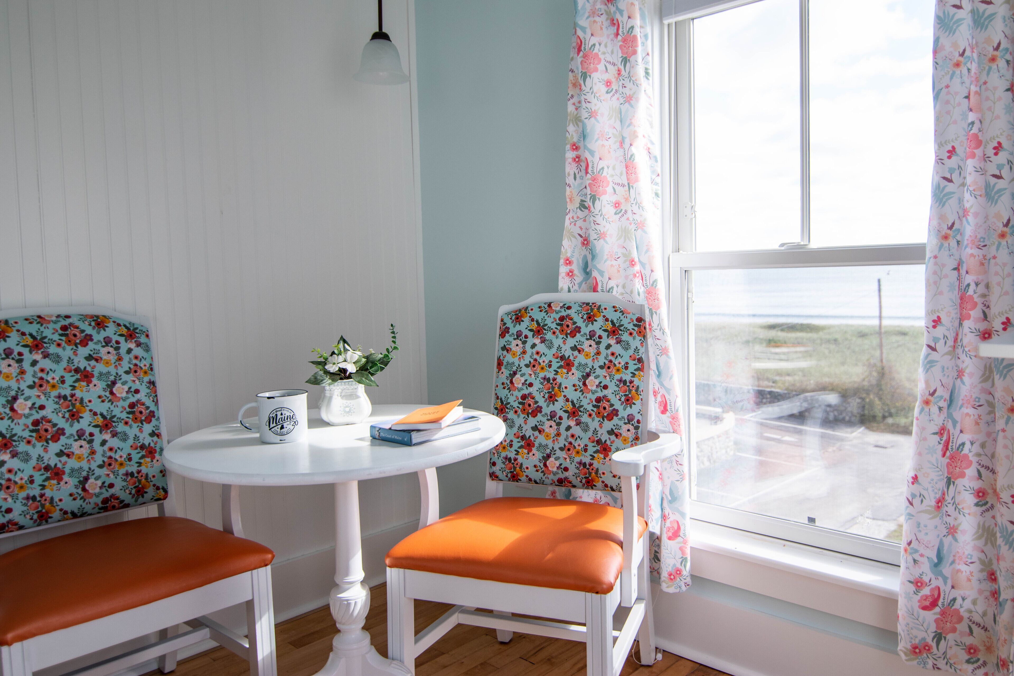 A room with two chairs with floral upholstery, a small round table with a white mug and a book, and a window with floral curtains.