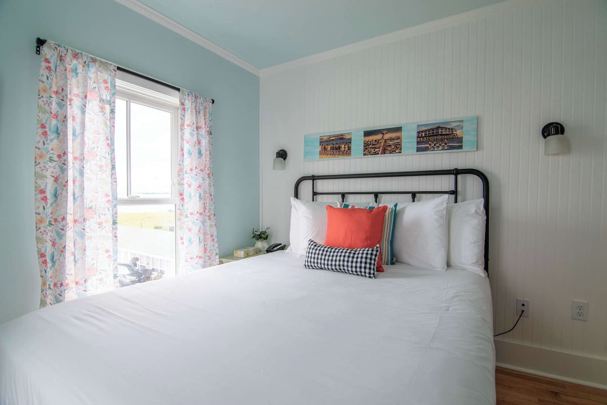 A bedroom with a bed, floral curtains, a window, wall-mounted artwork, and a nightstand with a lamp.