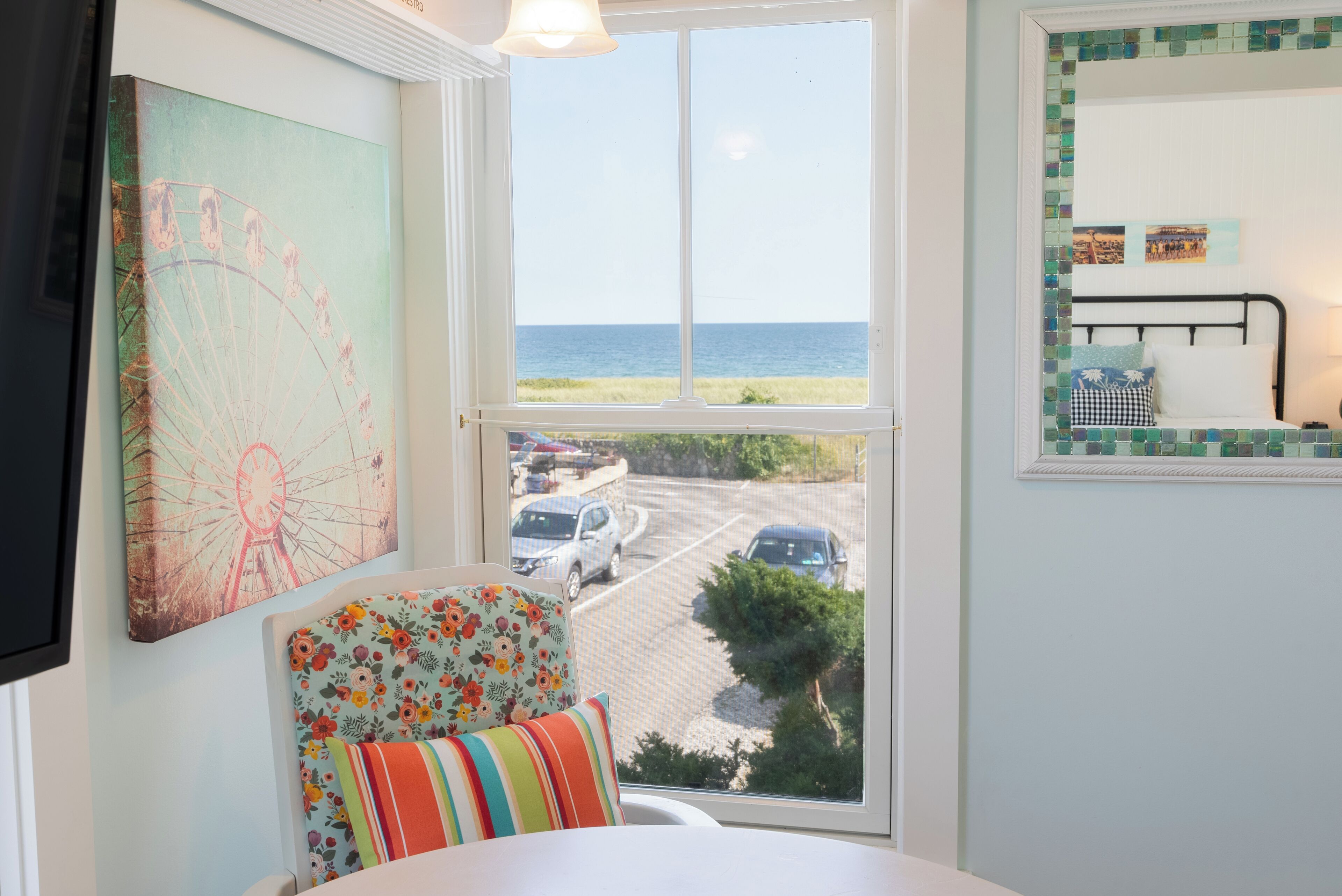 A room with a view of the sea, a chair with a floral cushion, and a bed with a checkered pillow.