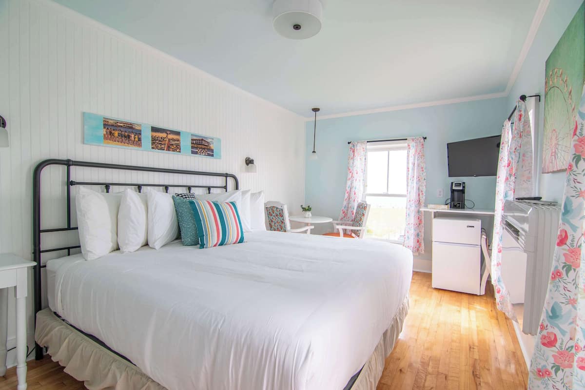 A bedroom with a large bed, a desk with a computer, a chair, and a small refrigerator.