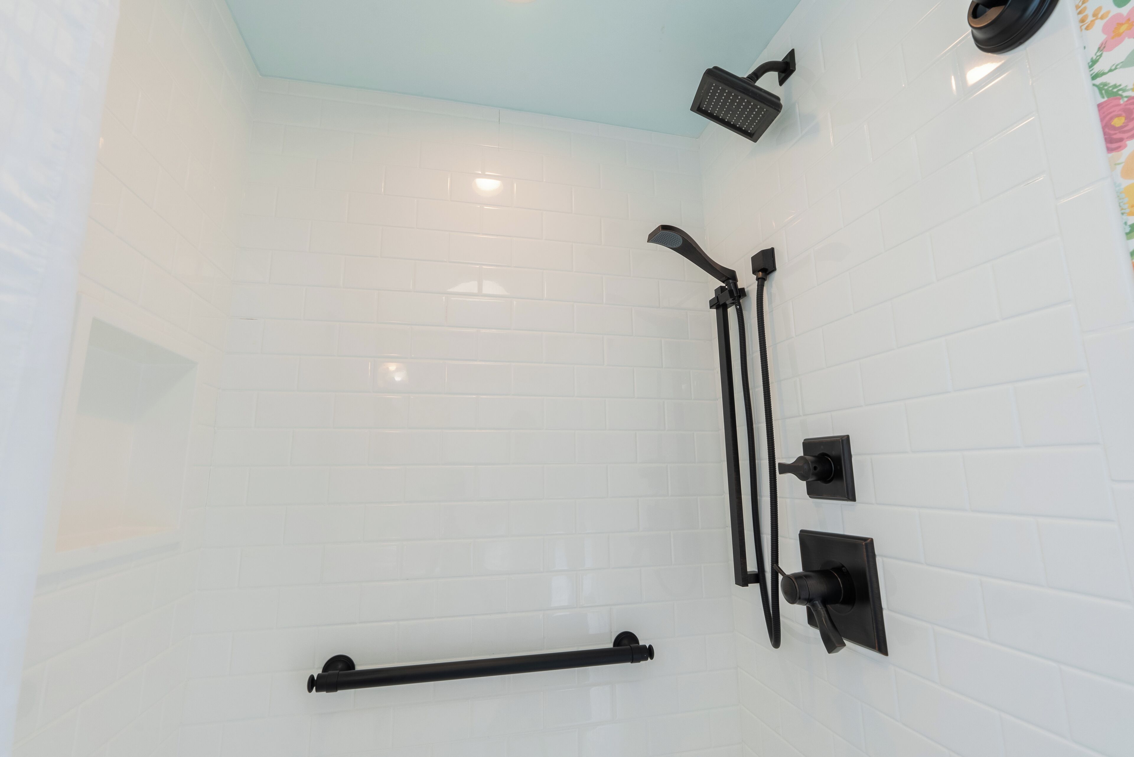 A bathroom with white tiled walls, a black towel rack, a black showerhead, and a black handheld showerhead.