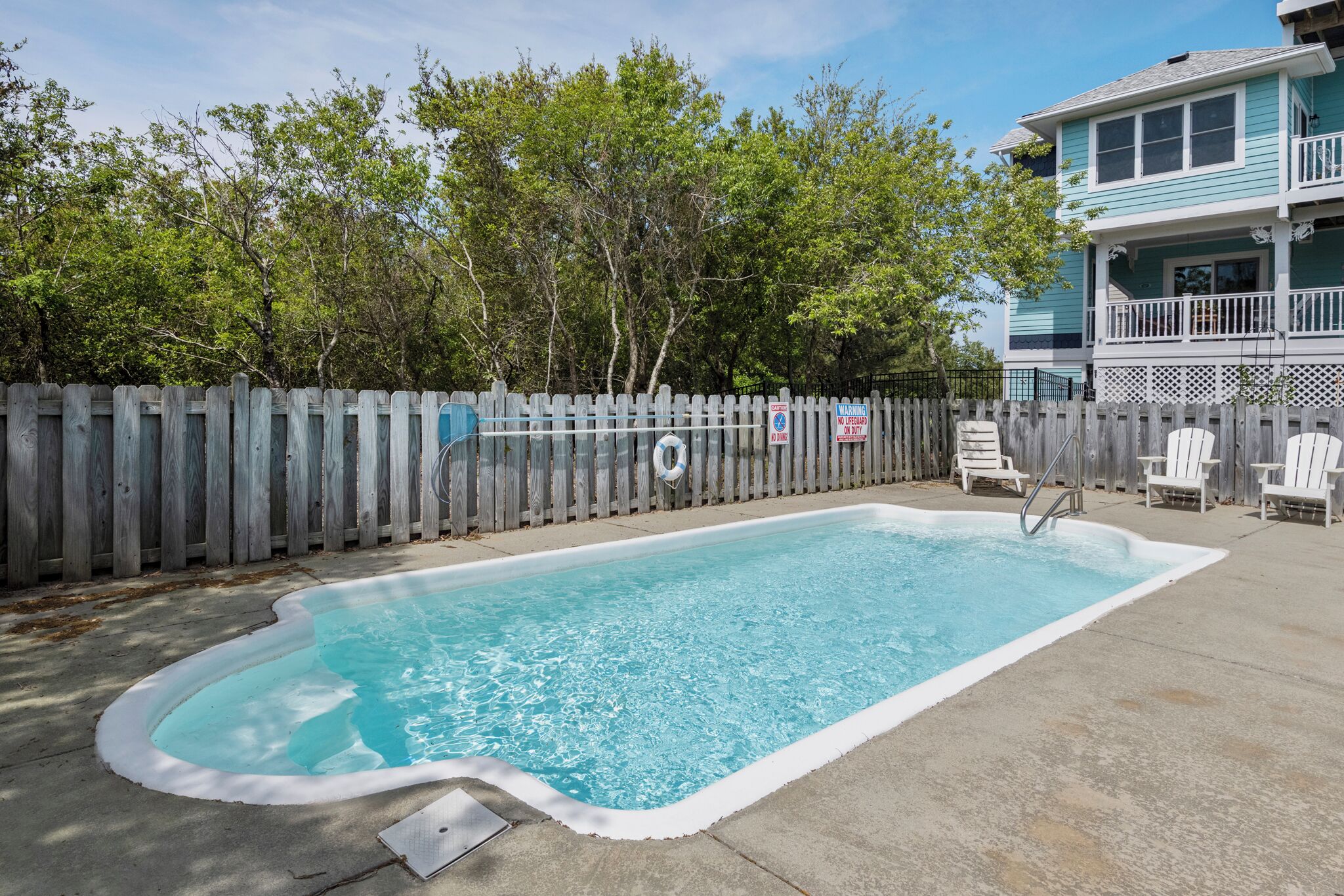 Une piscine rectangulaire avec un bord blanc, entourée d’une clôture en bois. On y trouve des chaises blanches et un escalier qui descend dans la piscine.