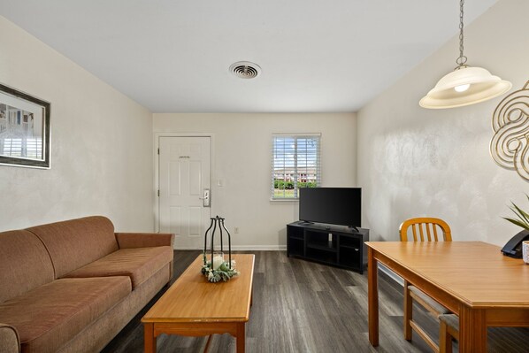 A living room with a brown sofa, a wooden table, a TV stand, and a dining table with chairs.