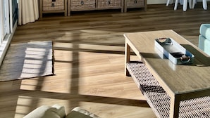 Una sala de estar con televisor, estanterías de madera, cestas de mimbre, una mesa de comedor con sillas y un ventanal con cortinas.