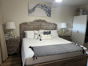 A neatly made bed with a patterned throw, two bedside lamps, a star-shaped nightstand, a wooden headboard, a framed artwork on the wall, and a wardrobe.