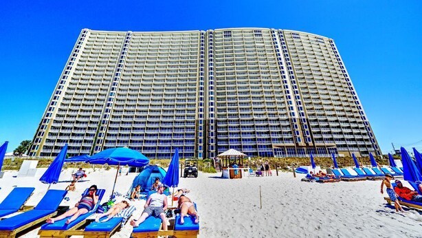 Un hotel grande con varios pisos y balcones, una playa arenosa al frente con tumbonas y sombrillas, y un cielo azul despejado.