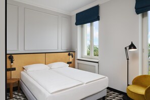 Et hotelværelse med seng, stol, et vindue med gardiner, en radiator og en lampe monteret på væggen.