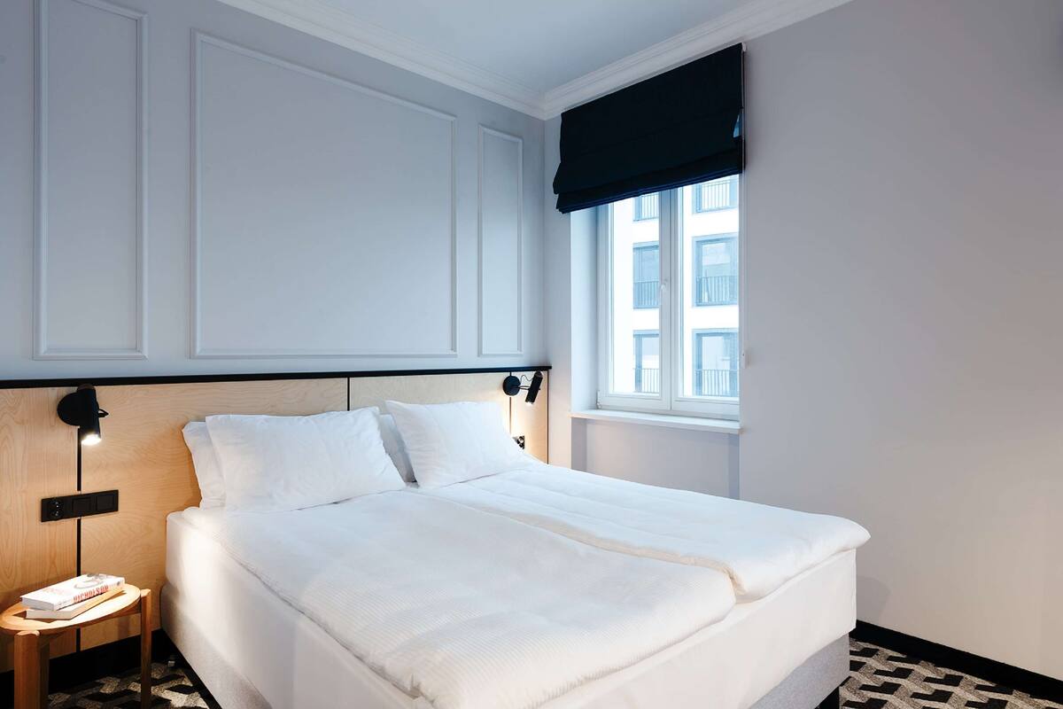 A neatly made bed in a hotel room with a headboard, a bedside table with a lamp, and a window with a dark blind.