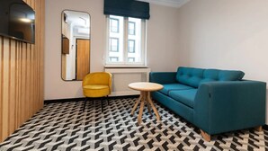 Quarto com piso estampado, um sofá azul, uma poltrona amarela, uma pequena mesa redonda e um espelho na parede.