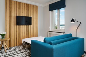 Una habitación con una cama, un televisor fijado a una pared de madera, una ventana con vistas, una lámpara y una mesita con un jarrón.