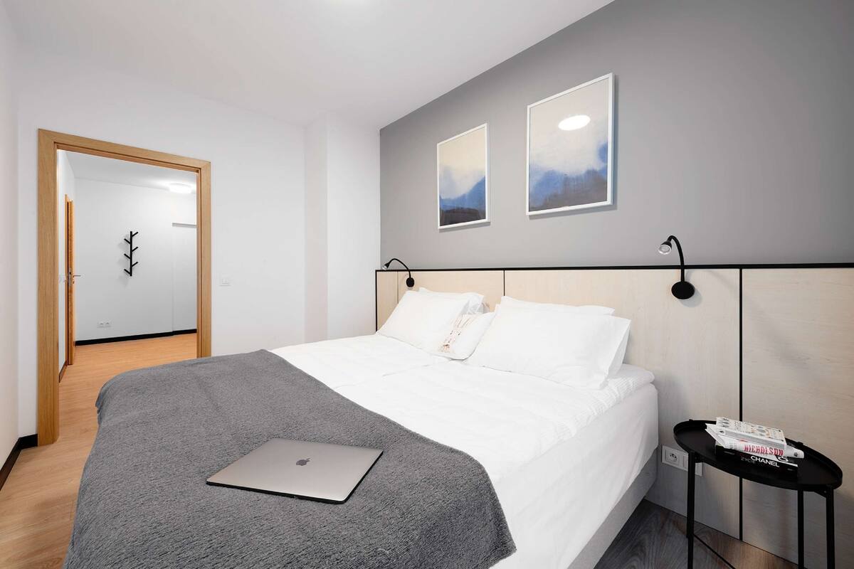 A modern hotel room with a neatly made bed, a laptop on the bed, a small side table with a remote control, and a door leading to another room.