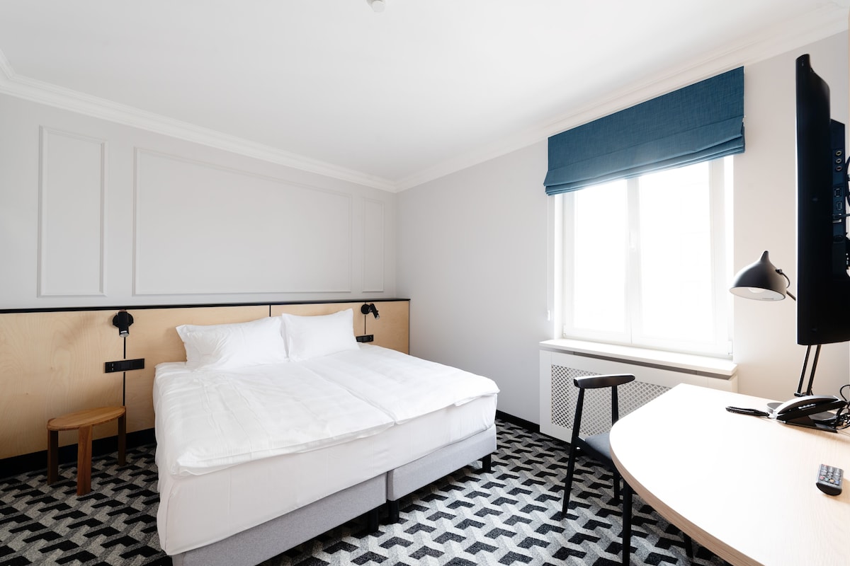 Quarto de hotel com cama, mesa de cabeceira, cadeira e escrivaninha com telefone.