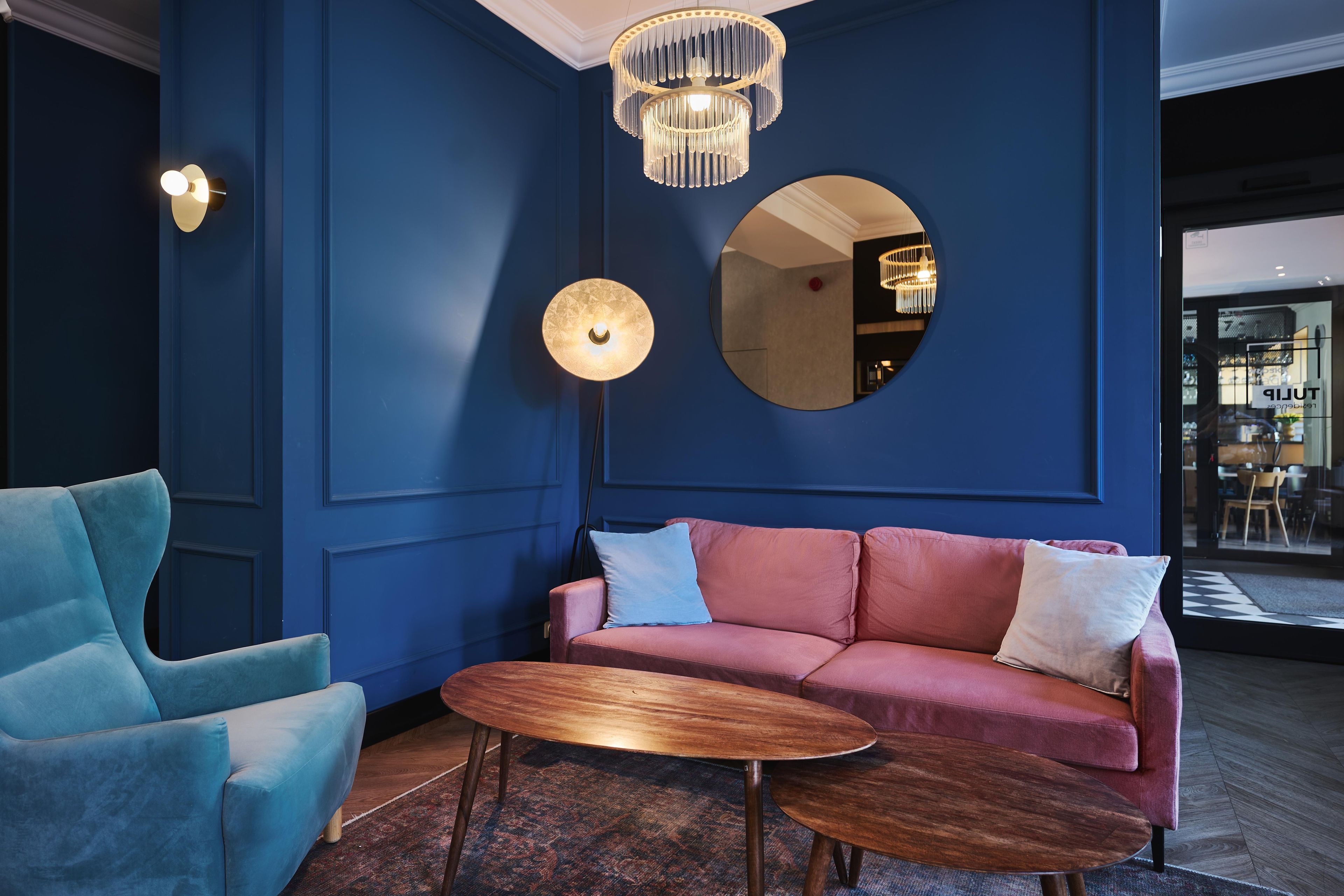 A room with a blue wall, a round mirror, a pink sofa with cushions, a blue armchair, and a wooden coffee table.