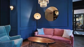 A room with a blue wall, a round mirror, a pink sofa with cushions, a blue armchair, and a wooden coffee table.
