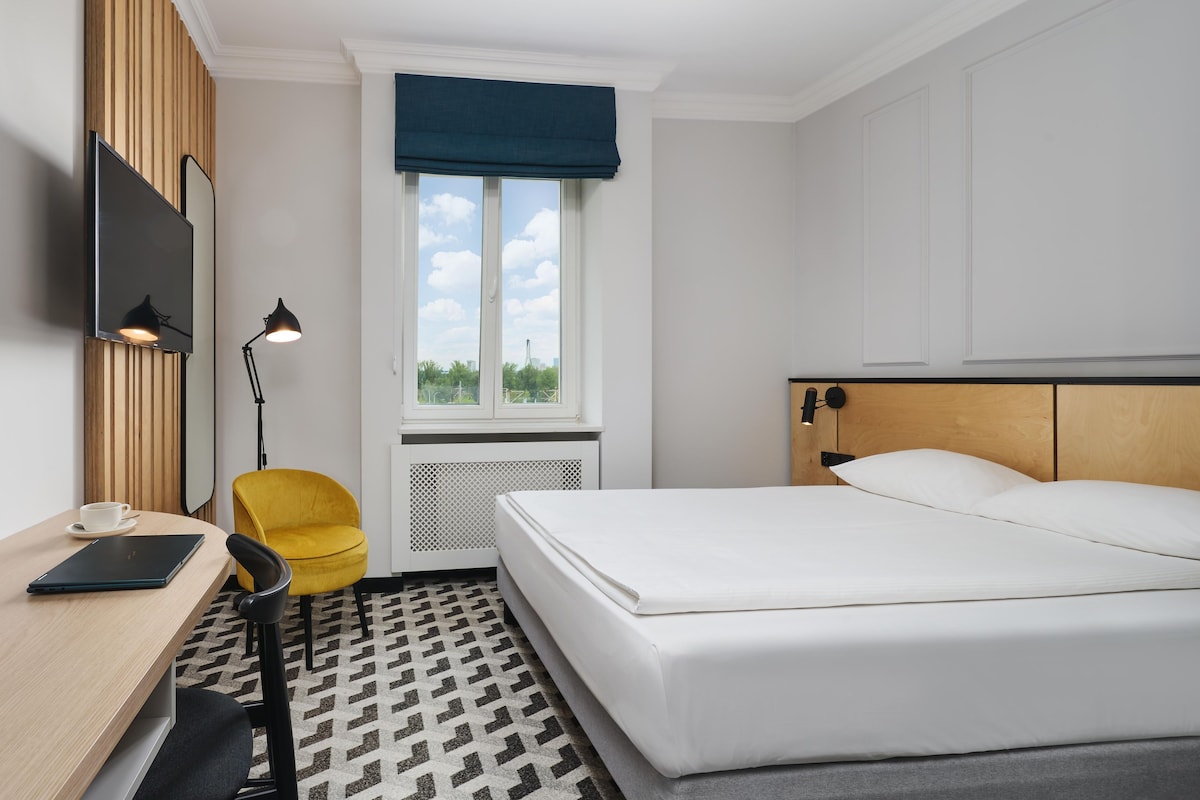Quarto de hotel com cama, escrivaninha com laptop, cadeira, TV fixada na parede e janela com vista para o exterior.
