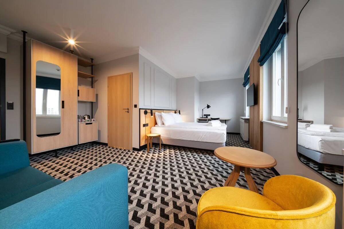 A hotel room with a bed, a desk, a chair, a mirror, and a patterned floor.
