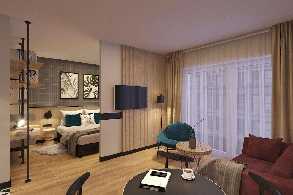 Quarto de hotel moderno com cama, televisão, área de estar com mesa e uma janela grande com cortinas.