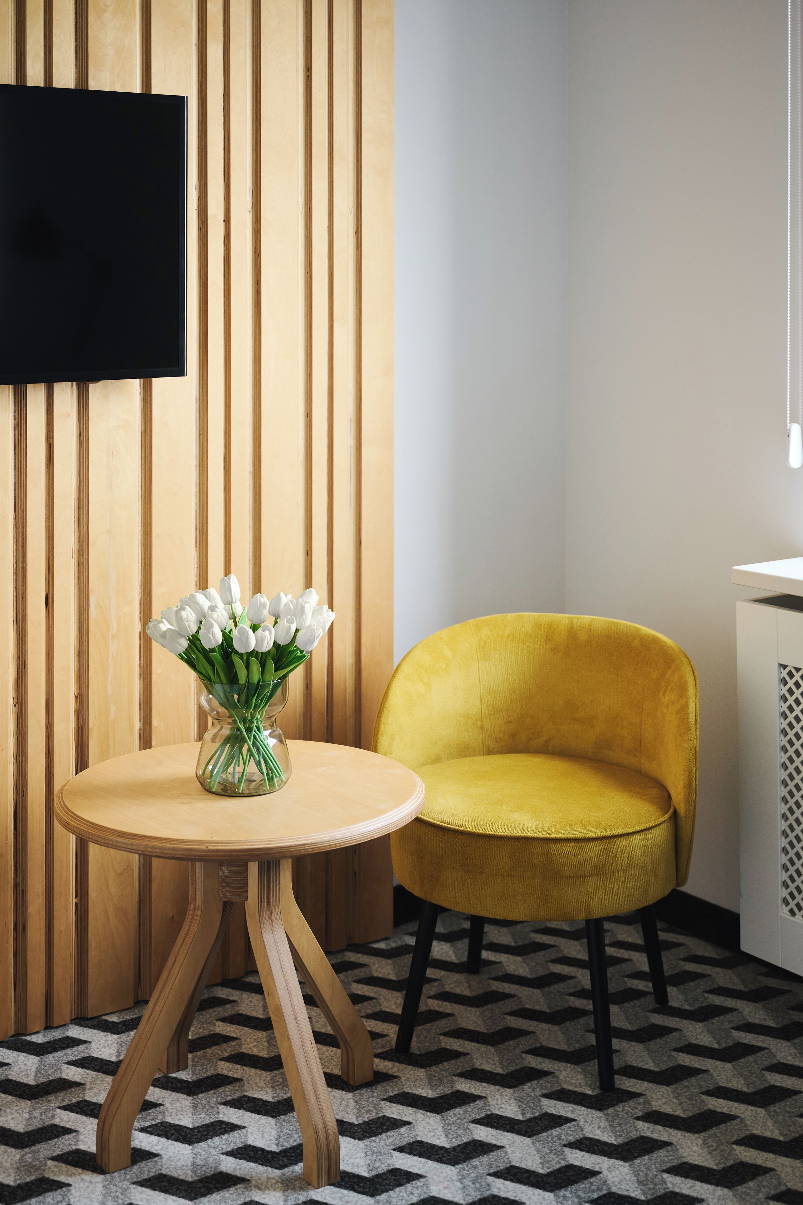 Een kamer met een wand van houten latten, een ronde houten tafel met een vaas witte bloemen, een gele fluweelstoel en een zwart-wit tapijt.