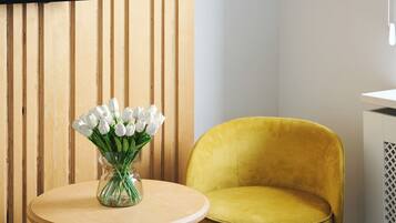 Een kamer met een wand van houten latten, een ronde houten tafel met een vaas witte bloemen, een gele fluweelstoel en een zwart-wit tapijt.