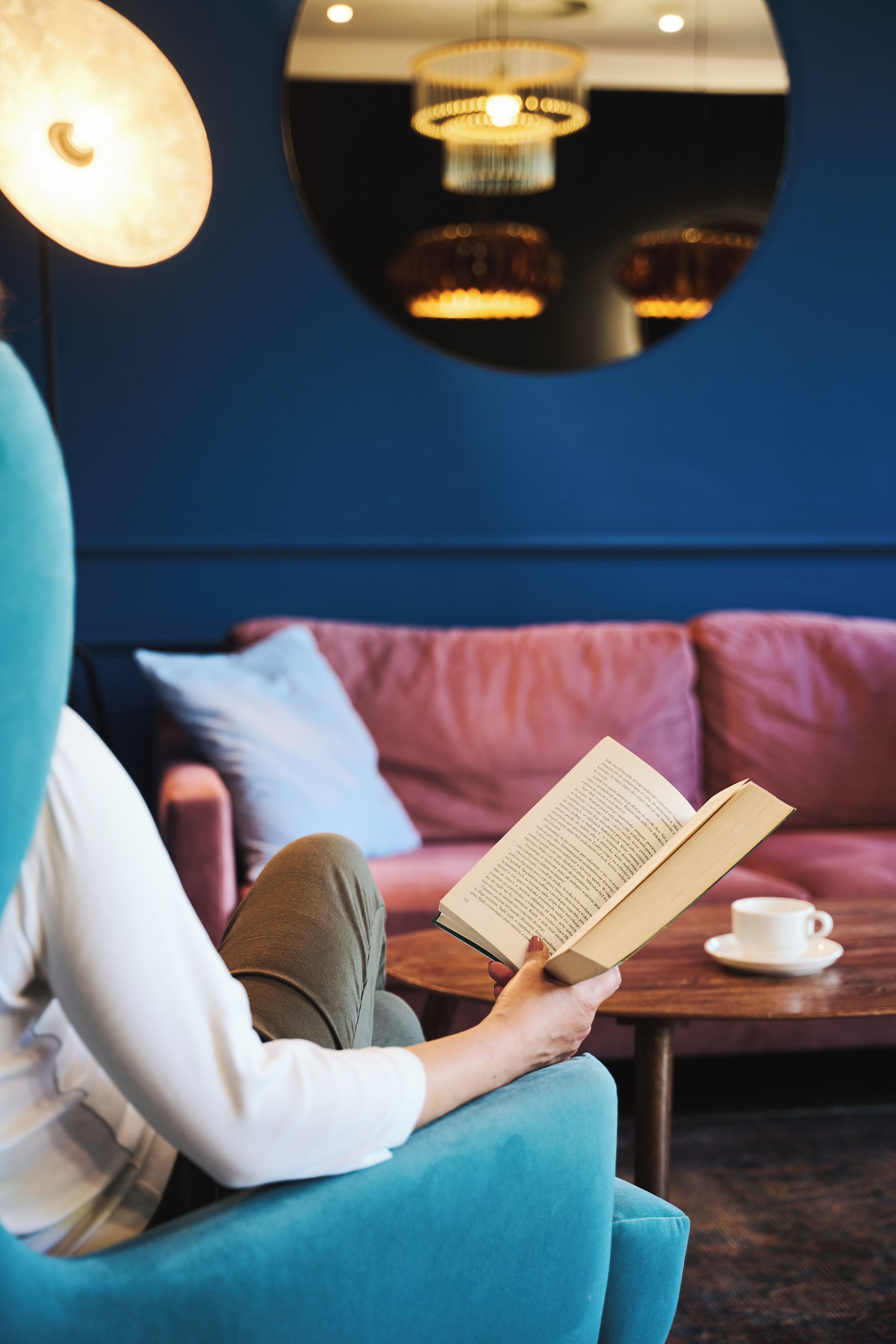 Una persona seduta su un divano color acquamarina, che legge un libro, con una tazza di caffè su un tavolino di legno di fronte.