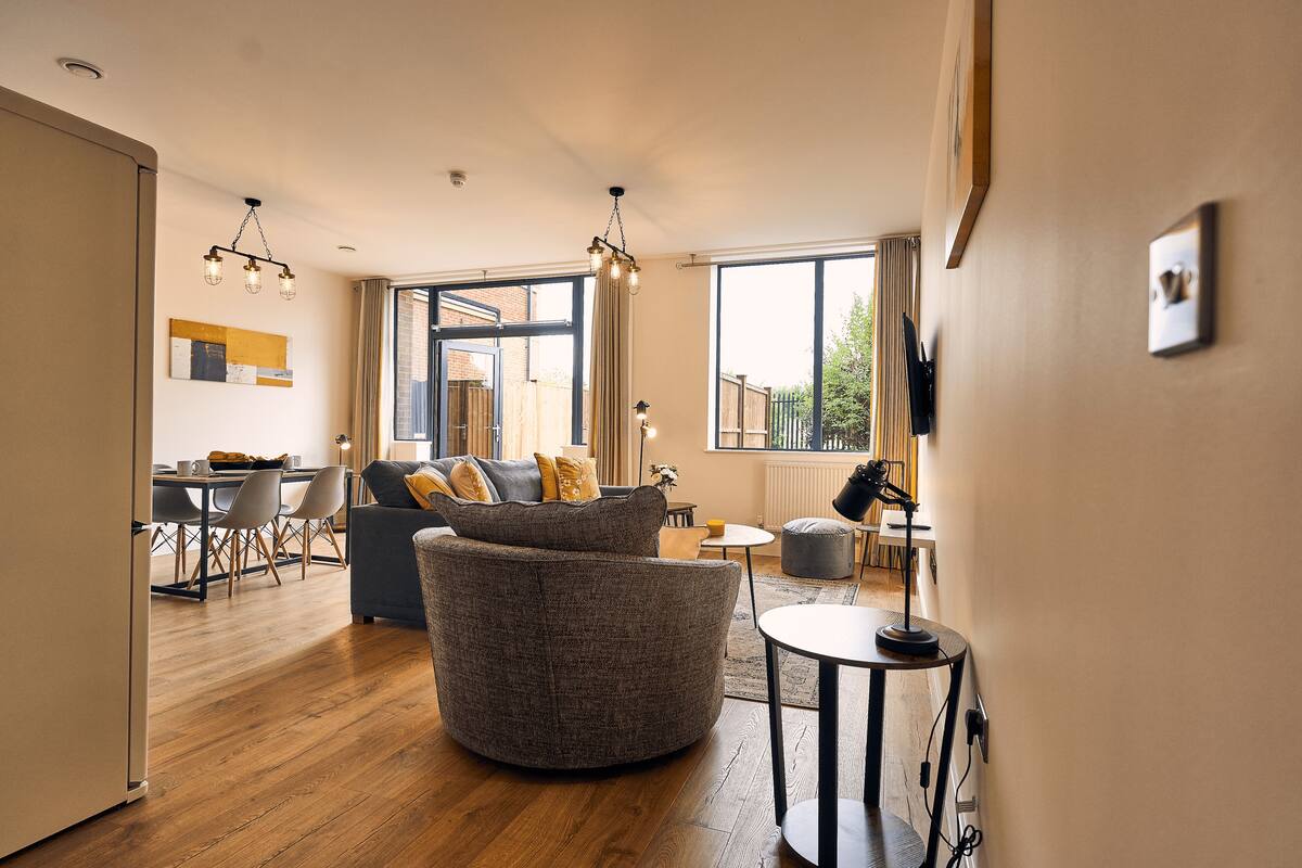 A modern living room with a round grey sofa, a dining area with a table and chairs, and large windows offering a view of the outdoors.