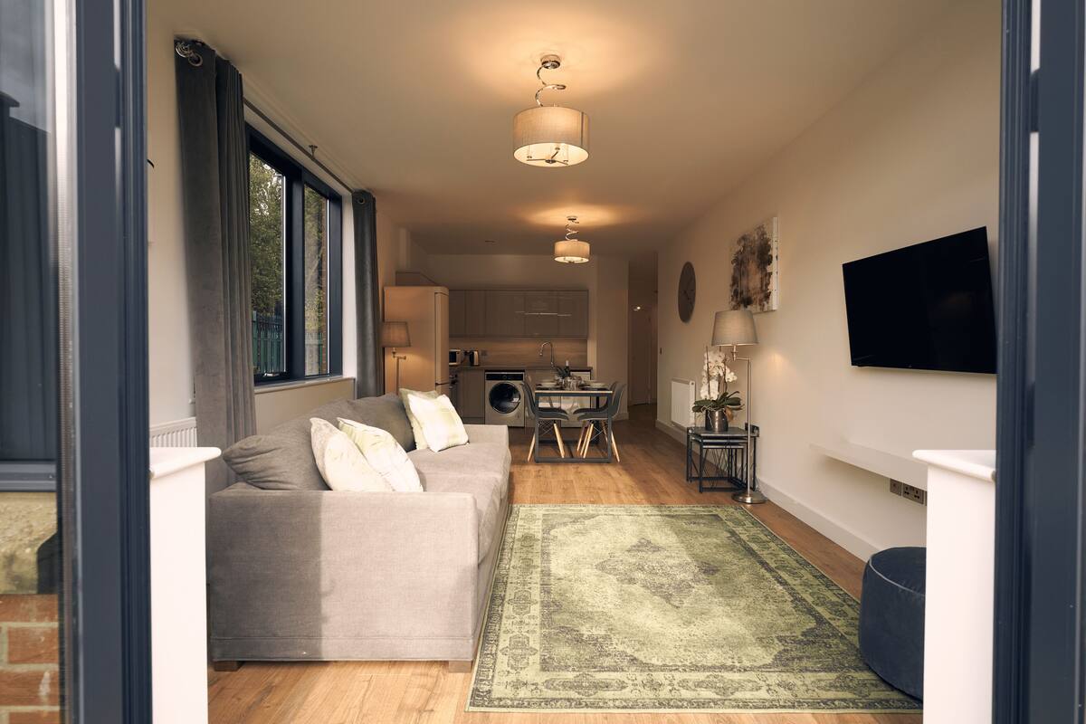 A modern living room with a grey sofa, a green rug, and a kitchen in the background.
