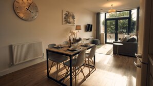 Comedor con mesa de madera, sillas y un reloj grande en la pared. Hay un radiador en la pared y una ventana con vistas a la vegetación exterior.